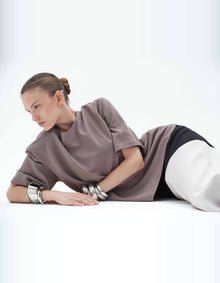 Reclining Model Wearing The Elamins Oversize T-Shirt In Taupe, Showing The Relaxed Silhouette, Wide Short Sleeves And Soft Fabric Folds.