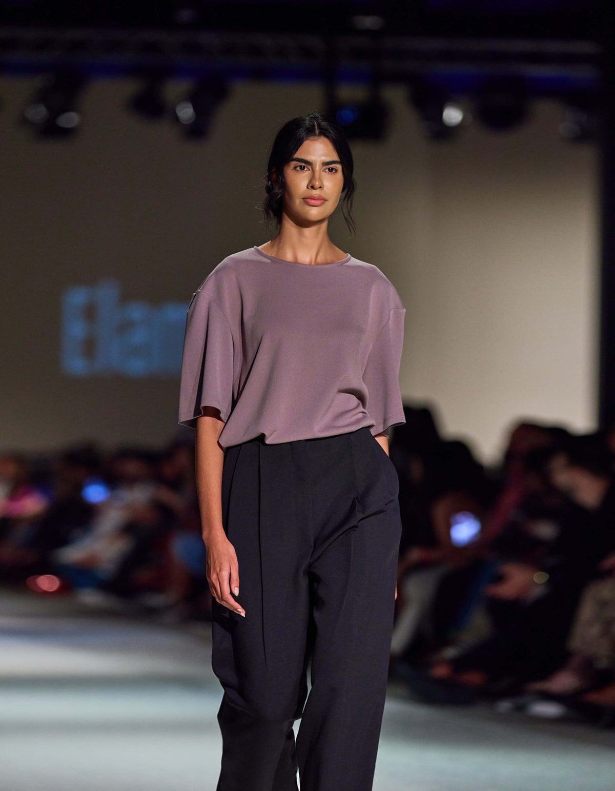 Side-Angle Runway View Of The Elamins Oversize T-Shirt In Taupe, Highlighting Its Fluid Drape And Oversized Shape, Paired With The Elamins Black Pleated Trousers At TheEventDXB.